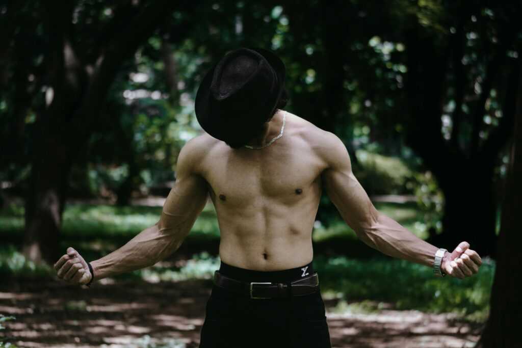 A shirtless man wearing a hat poses confidently outdoors in a forest setting.