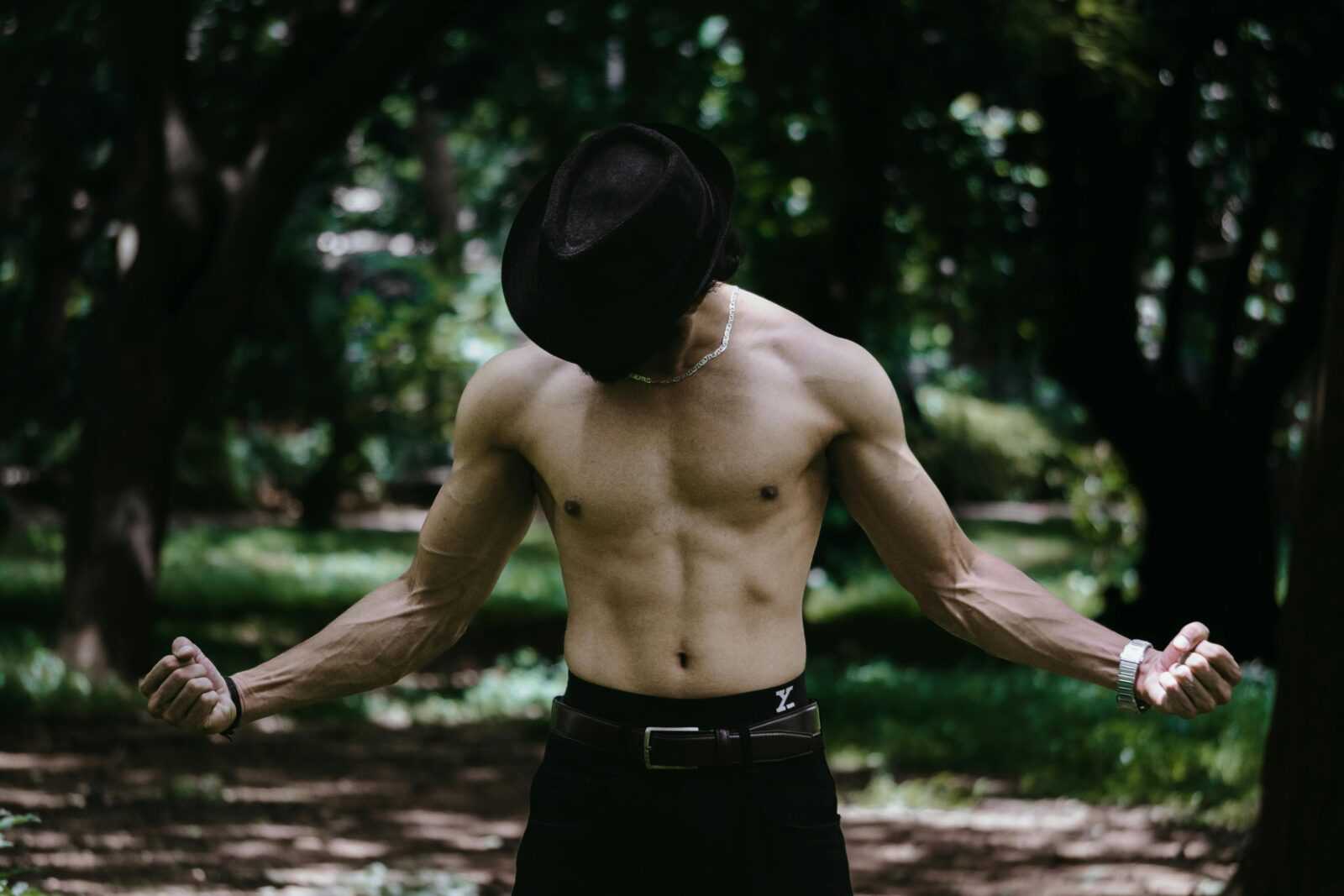 A shirtless man wearing a hat poses confidently outdoors in a forest setting.